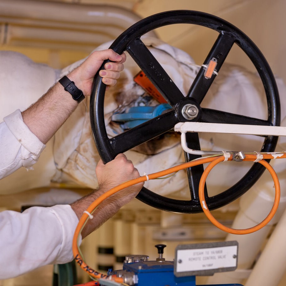 A person wearing a smartwatch turns a large black industrial valve wheel attached to insulated pipes, with an orange cable looped round the mechanism—a glimpse into P&O Cruises technical careers in action.
