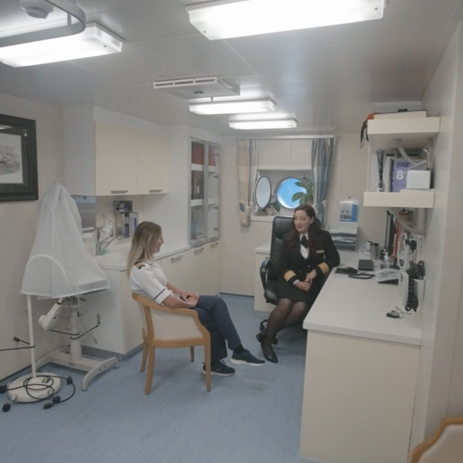Two women in uniforms are talking inside a clean, well-lit medical office on a ship. One woman sits at a desk with a computer, while the other is seated nearby. Medical equipment and books are visible in the room.