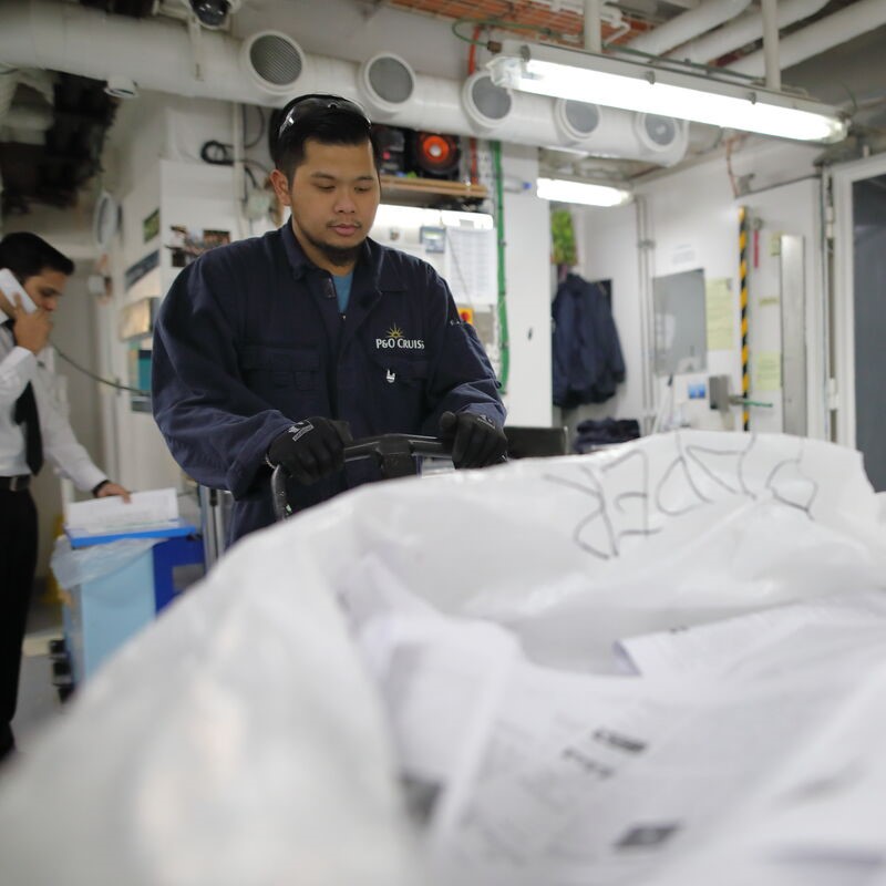 A man in a navy uniform and gloves, possibly from P&O Cruises technical careers, pushes a trolley with papers through a busy indoor workspace whilst another person in the background talks on the phone.