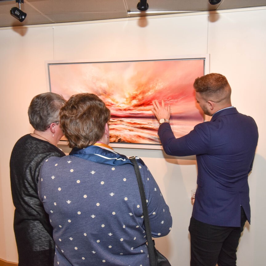 Three people stand in front of a colourful abstract painting in a gallery. One person gestures towards the artwork while the others observe closely, all with their backs to the camera.