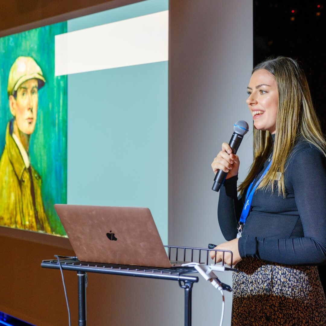 A woman holding a microphone stands beside a laptop, giving a presentation. A painting of a person in a cap is projected on the screen behind her.