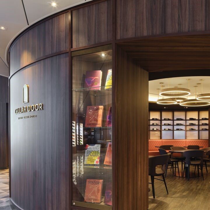 Curved wooden wall with a glass display of colourful books, sign reading “Cellar Door,” and a modern, warmly lit interior reminiscent of inviting cruise ship bar jobs, with round tables, chairs, and shelves in the background.