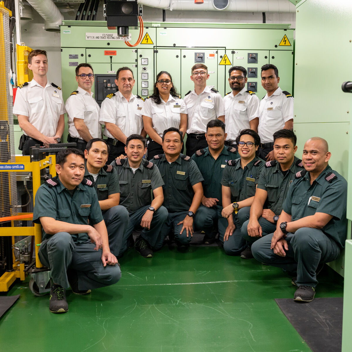 A group of 15 people in two different uniforms—white shirts and dark green shirts—pose and smile together in an industrial control room, showcasing the teamwork behind P&O Cruises technical careers amid machinery and control panels.