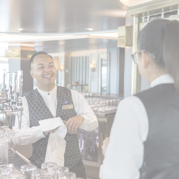 A smiling barman in a waistcoat wipes a glass behind the bar whilst talking to a colleague, who stands in the foreground with her back to the camera. The bar area is bright and modern.