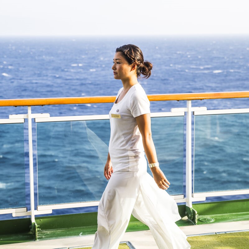A woman in a white outfit walks on the deck of a ship with the sea visible in the background on a sunny day.
