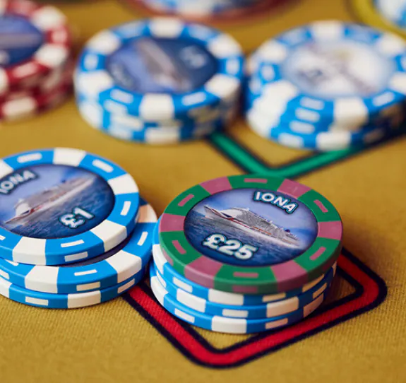 Stacks of colourful poker chips labelled IONA with denominations of £1 and £25, featuring an image of a cruise ship, placed on a yellow casino table with red and black markings.