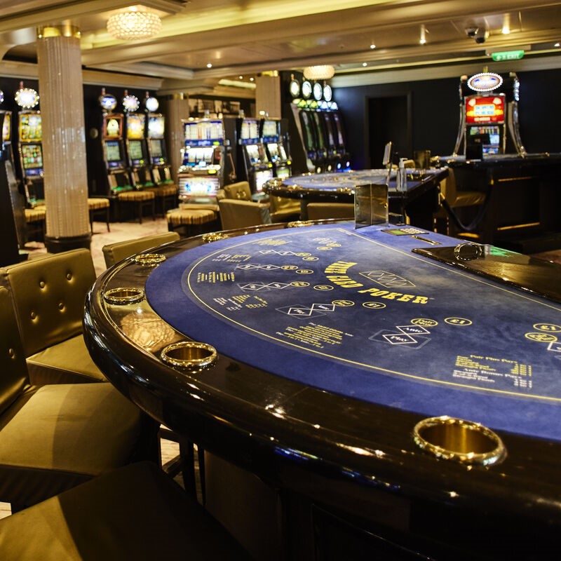 A casino interior with empty gaming tables in the foreground, upholstered chairs, and rows of fruit machines with bright screens in the background under warm lighting.