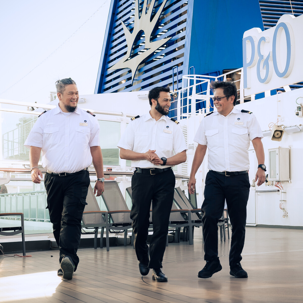Three men in white short-sleeve uniforms and black trousers walk together on the deck of a ship, smiling and talking. The P&O Cruises logo and blue funnel are visible in the background.