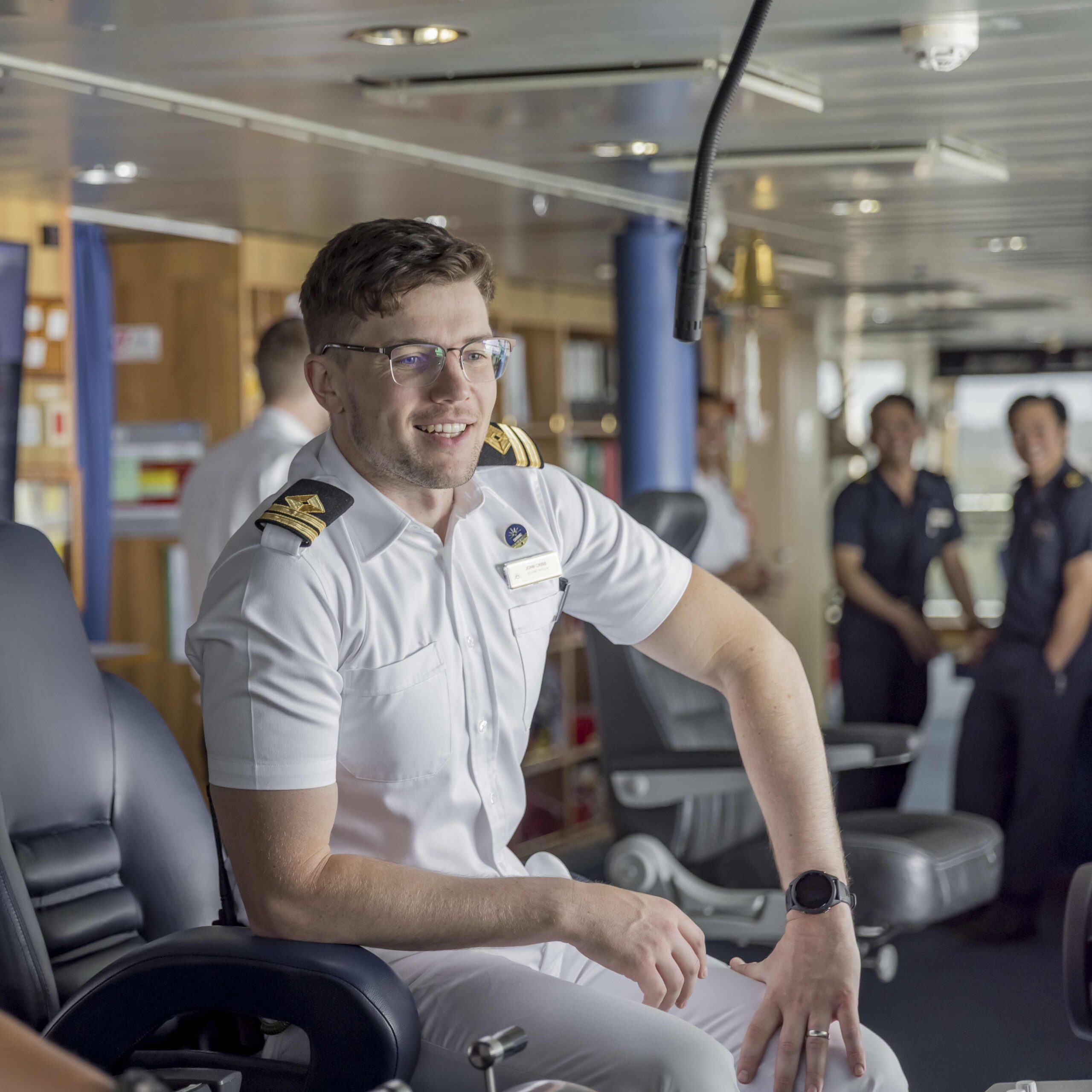 A smiling ship officer in a white uniform sits at a control station on the Bridge with three other crew members in uniform standing in the background, all appearing relaxed and engaged.
