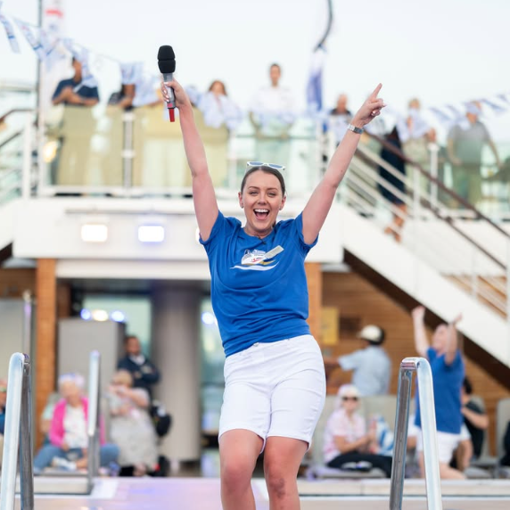 A smiling woman in a blue shirt and white shorts holds a microphone and raises both arms in excitement on a cruise ship deck, with people and festive decorations in the background.