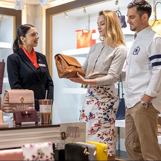 A woman examines a brown handbag in a shop while a man stands beside her. A shop assistant in a black blazer and red scarf is smiling and assisting them. Shelves of colourful handbags are displayed in the background.