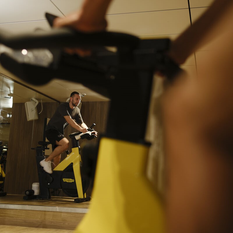A man wearing a black shirt and shorts rides a yellow exercise bike in a gym, with his reflection visible in the mirror behind him. The blurred foreground shows part of another exercise bike.