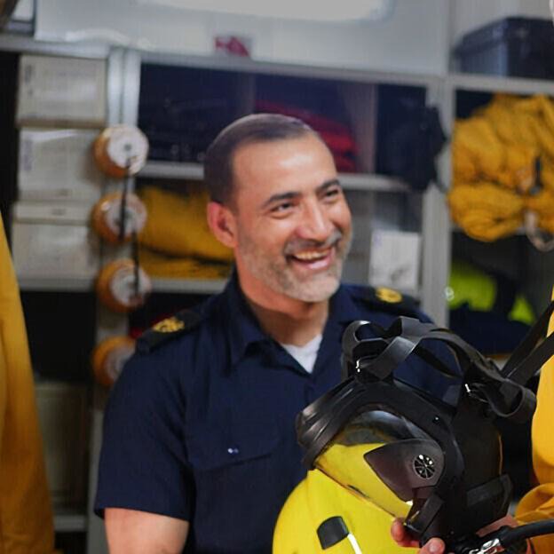 A man in a dark uniform smiles as he holds safety gear, showcasing P&O Cruises deck careers in a room with shelves containing yellow equipment and coiled hoses.