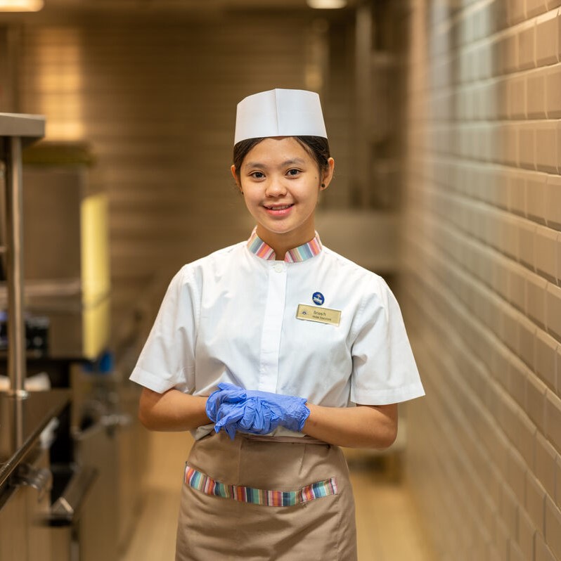 A smiling catering worker stands in a kitchen corridor, wearing a white uniform, apron, hat, and blue gloves—typical attire for cruise ship bar jobs—with hands clasped and a name badge visible. The background features tiled walls and stainless steel equipment.
