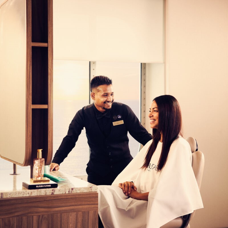 A smiling hairdresser in a black uniform stands beside a seated woman with long straight hair in a salon chair; both appear happy. The salon has a large mirror, and sunlight comes through a window in the background.