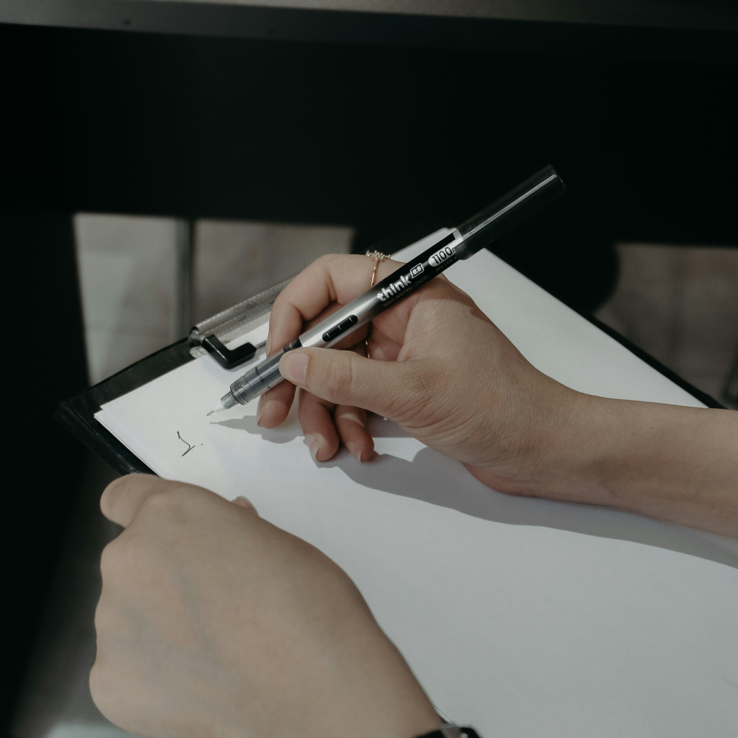 A person holding a pen writes on a blank sheet of paper attached to a clipboard, with only a small mark visible on the page.