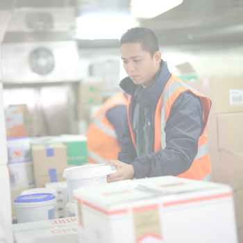 A worker wearing a reflective safety vest handles a white container in a storeroom filled with boxes and other packages.