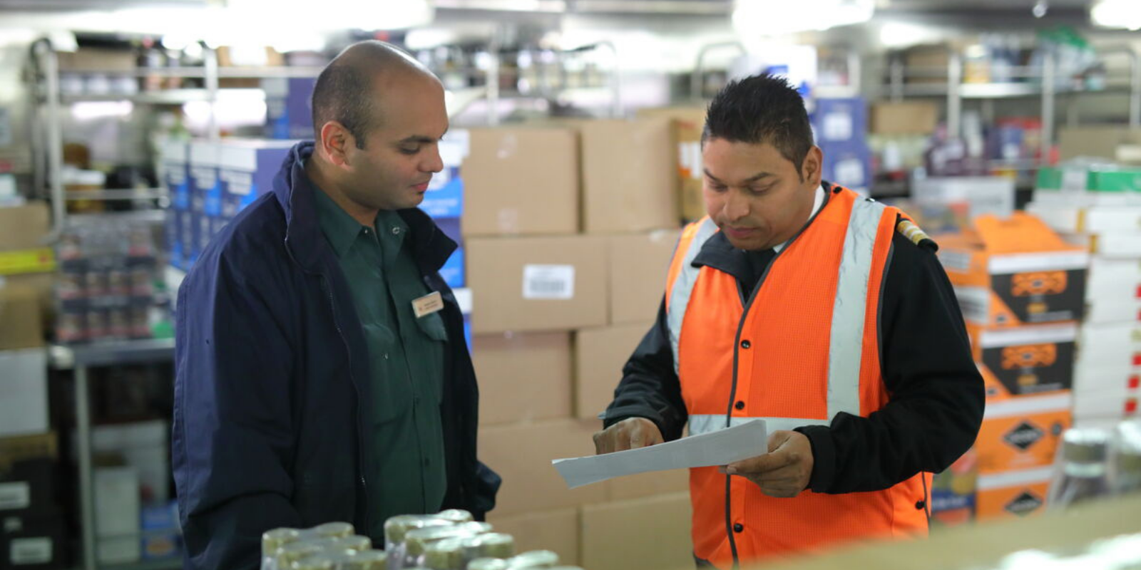 Two men stand in a warehouse among shelves and boxes; one in a dark jacket listens whilst the other, wearing an orange safety vest, points to a sheet of paper they are both reviewing.