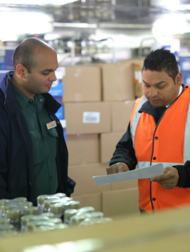 Two men stand in a warehouse; one wears a green shirt and name badge, the other a bright orange safety vest. They review paperwork together near shelves of goods and stacked cardboard boxes.