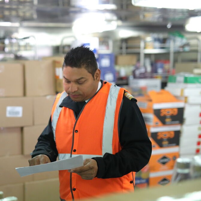 A man wearing an orange safety vest holds and reviews a sheet of paper in a warehouse filled with stacked boxes and various supplies.