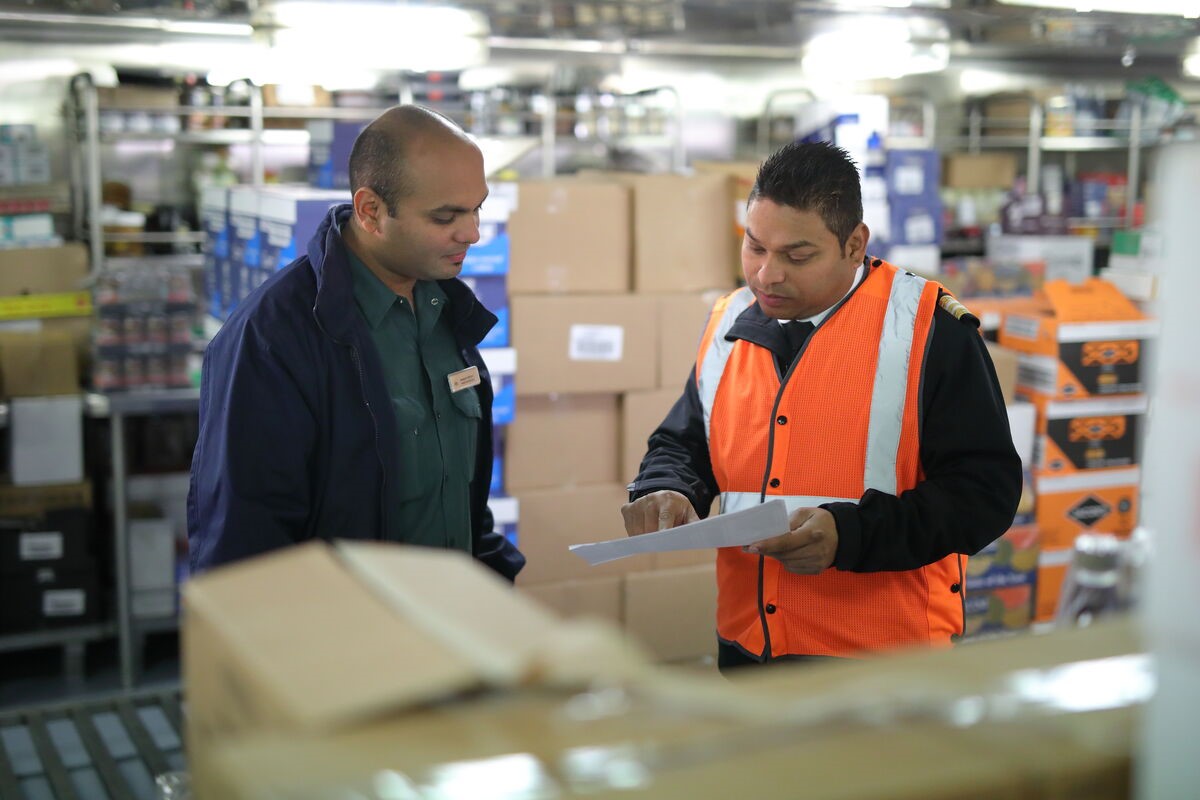 Two workers in a warehouse, one wearing an orange safety vest and holding a clipboard, discuss paperwork surrounded by shelves and boxes—quite different from the lively atmosphere of cruise ship bar jobs.