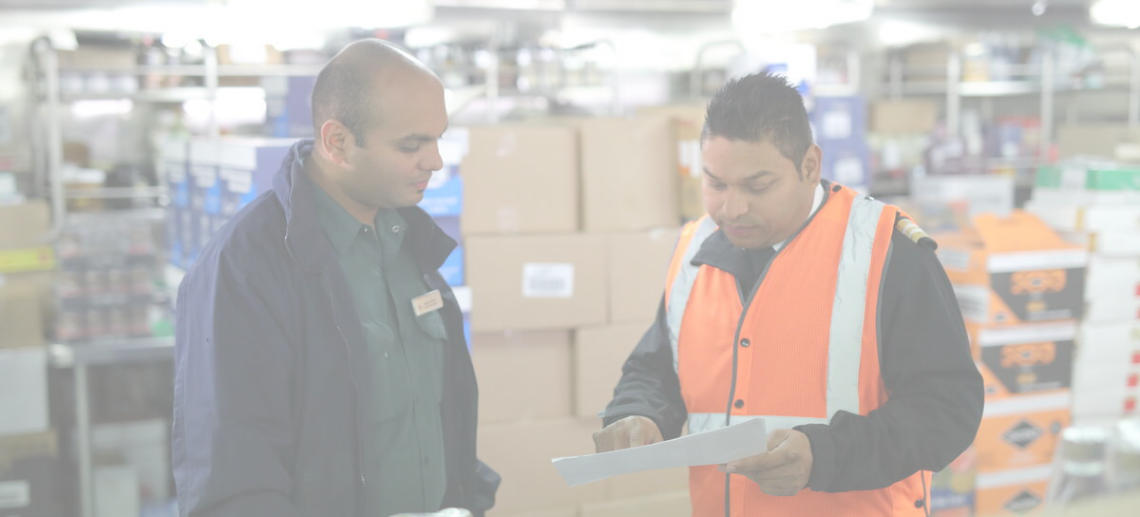 Two men stand in a warehouse, surrounded by boxes and shelves. One man in a green shirt listens whilst the other, in an orange safety vest, points at a document they are both examining.