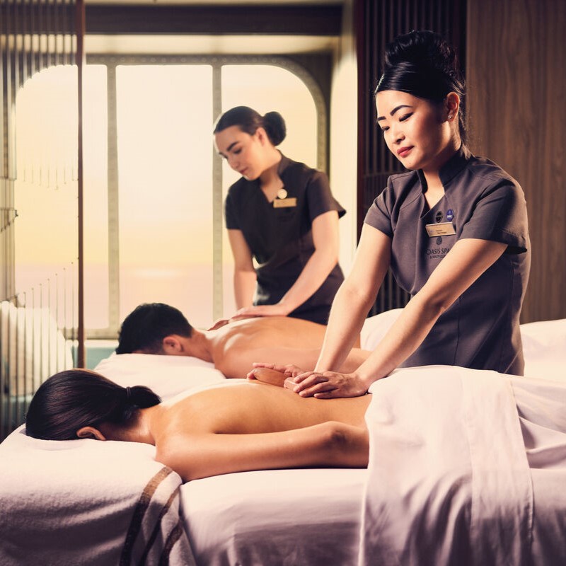 Two massage therapists in uniform are giving back massages to a man and a woman lying face down on massage tables in a spa room with soft lighting and a calm atmosphere.