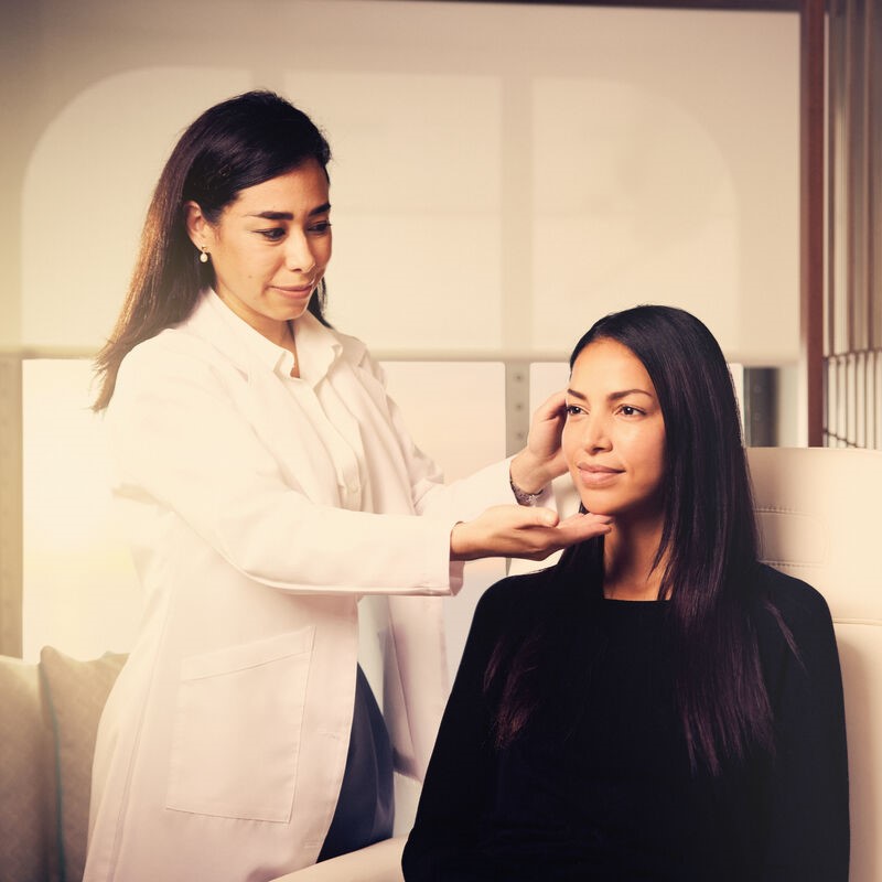 A female doctor in a white coat gently examines the jawline of a seated woman with long dark hair in a modern, well-lit medical surgery.
