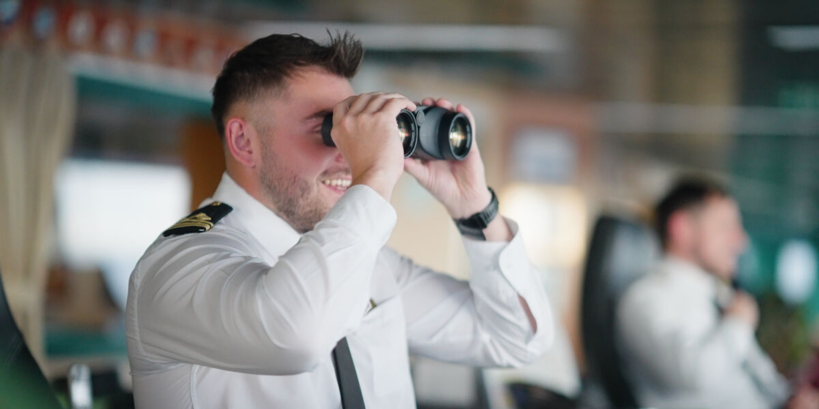 A man in a uniform smiles as he looks through binoculars indoors, showcasing the focus and teamwork found in P&O Cruises deck careers, whilst another uniformed person is blurred in the background.