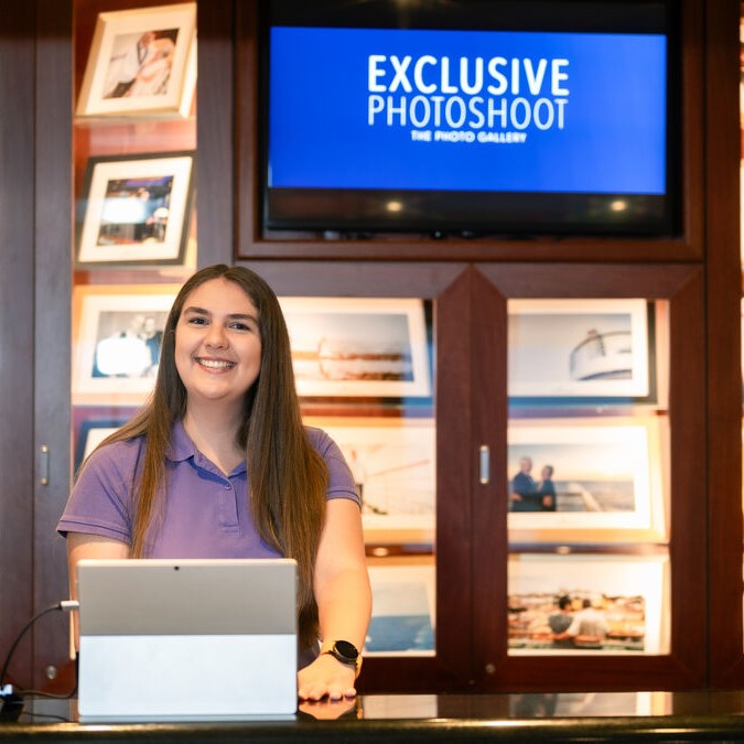 A smiling woman with long brown hair and a purple polo shirt stands behind a counter with a tablet. Behind her are framed photos and a screen displaying EXCLUSIVE PHOTOSHOOT: THE PHOTO GALLERY.
