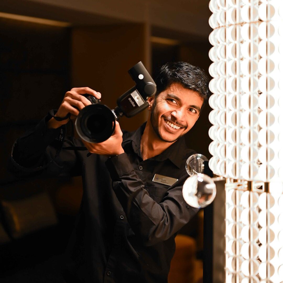 A smiling man in a black shirt holds a professional camera with a flash, standing beside a bright, decorative light fitting. The background is dimly lit, highlighting the subject and the light.
