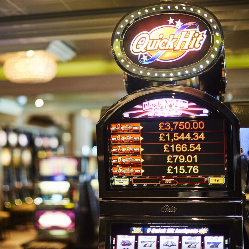 A close-up of a Quick Hit fruit machine in a casino, displaying potential winnings in pounds sterling, with blurred fruit machines and lights visible in the background.