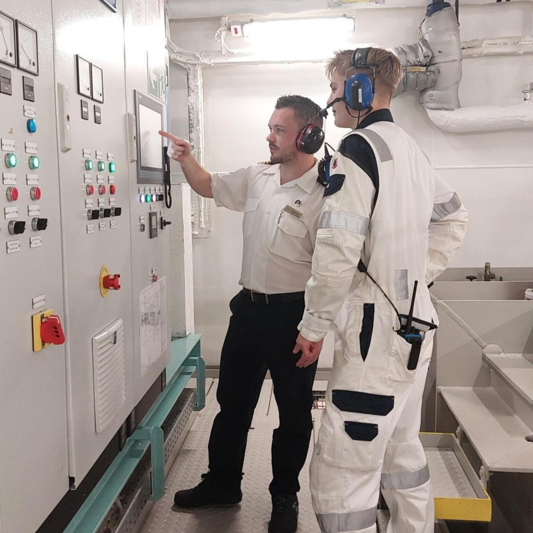 Two people in work uniforms and headsets stand in front of a control panel, one pointing whilst explaining something to the other—a glimpse into P&O Cruises technical careers in a high-tech, dynamic environment.