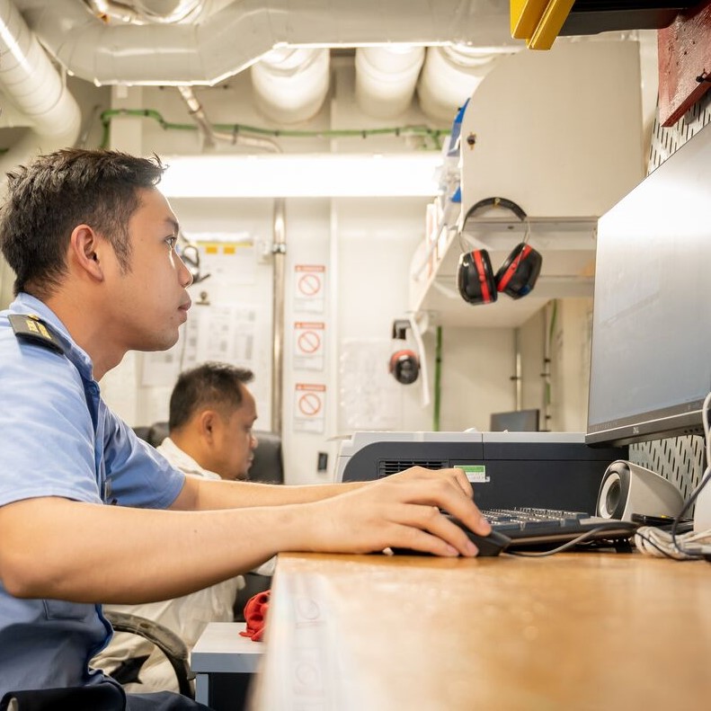 Two people in uniform work at computer stations in an industrial-looking office with equipment, headsets, and warning signs visible. One person is focused on a monitor—showcasing the dynamic environment of P&O Cruises technical careers.