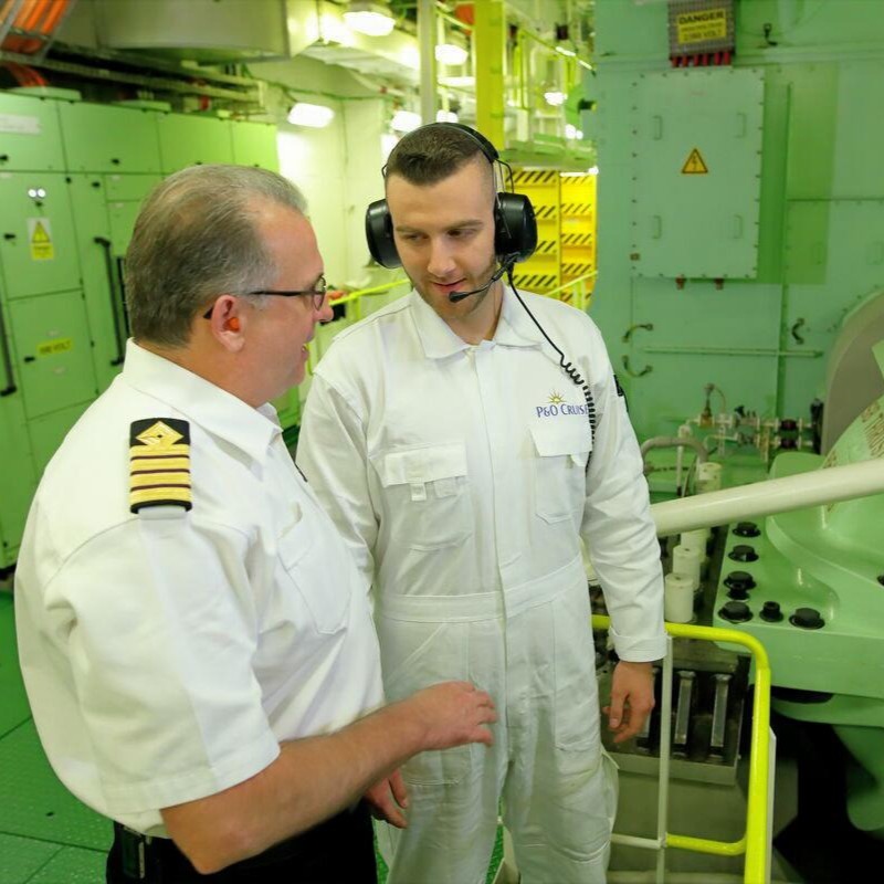 Two men in uniforms stand in an industrial engine room; one wears a headset and white overalls labelled “P&O Cruises,” while the other, in a staff chief's shirt with stripes, speaks to him. The background features green machinery and equipment.