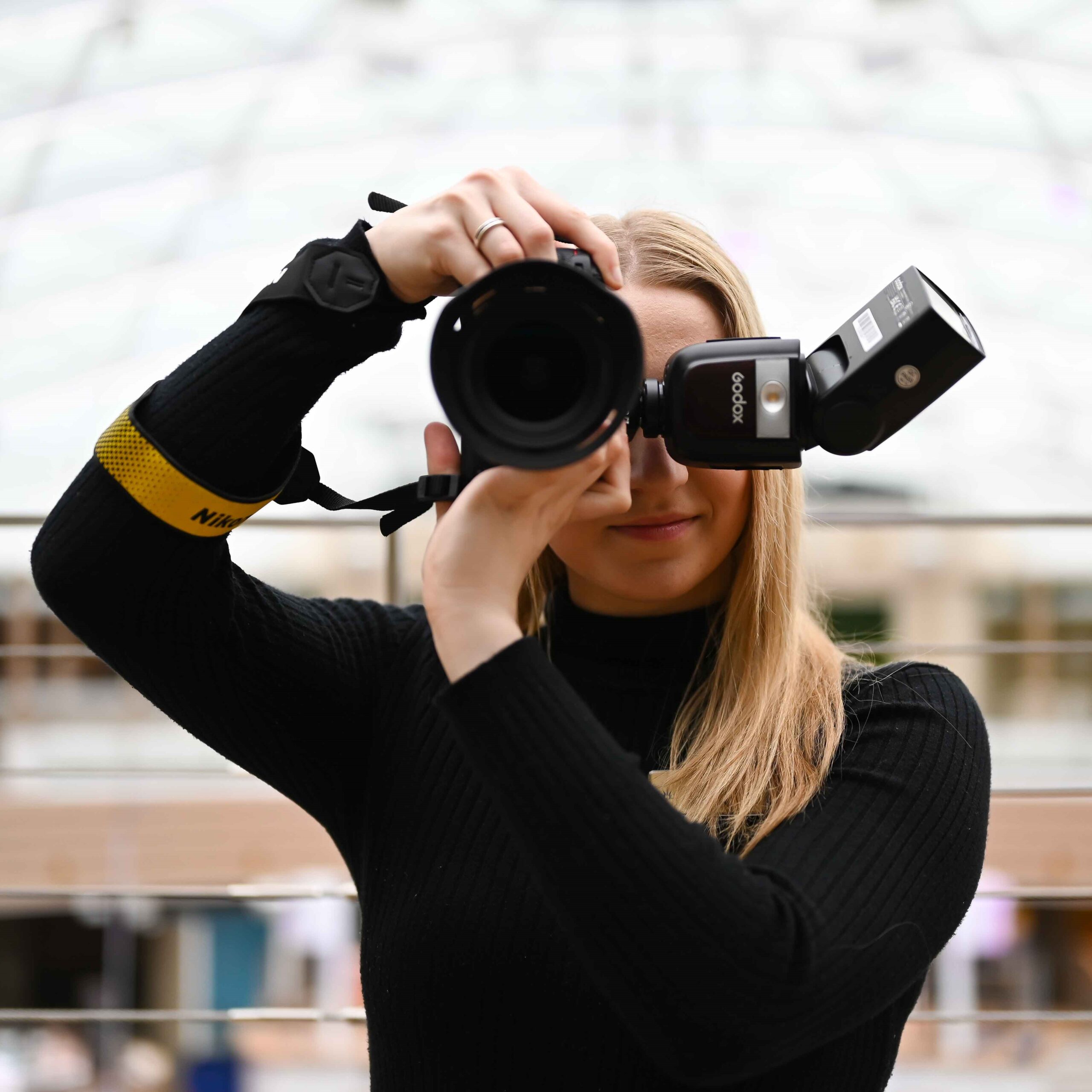A person with long blonde hair and a black polo neck holds a professional camera up to their face, preparing to take a photo. The background is blurred, and they wear a yellow camera strap on their wrist.