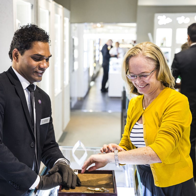 A smiling woman in a yellow cardigan tries on a bracelet at a jewellery shop counter, assisted by a male employee in a suit and gloves. Jewellery pieces are displayed on a tray between them.