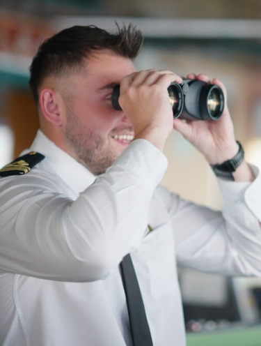 A man in a white uniform with black shoulder epaulettes looks through binoculars, smiling. He appears to be in a professional setting, possibly exploring P&O Cruises deck careers on a ship or in a control room.