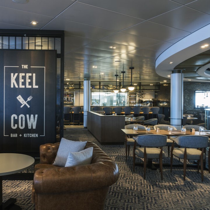 Modern restaurant interior with leather and fabric seating, wooden tables, and stylish lighting. A large sign reads The Keel Cow Bar + Kitchen near the entrance—a perfect setting reminiscent of upmarket cruise ship restaurant jobs. The bar area is visible in the background.