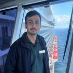 Crew member in a dark work uniform standing on the ship's bridge with railings and the sea in the background.