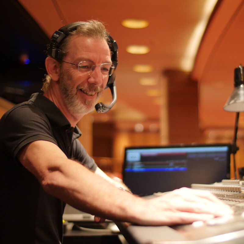 Crew member wearing a headset working inside a control or audio room, with equipment, screens, and warm ambient lighting in the background.