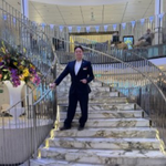 Crew member standing on a ship’s upper deck overlooking a multi-level interior atrium with balconies, glass, and decorative features.