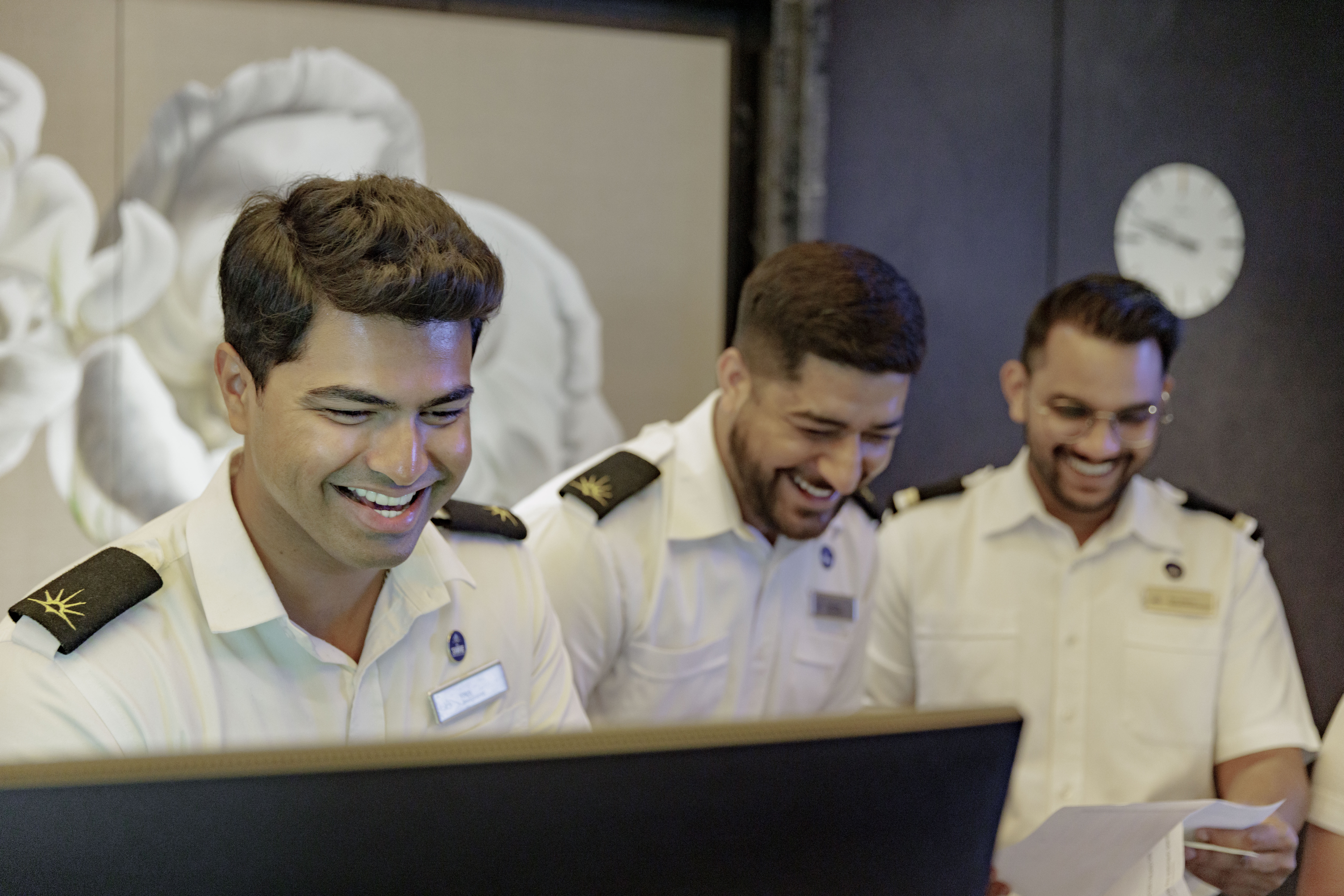 Three smiling men in white uniforms stand behind a desk, looking at a computer screen and a piece of paper, appearing to share a light-hearted moment at work. A clock and flower artwork are visible in the background.