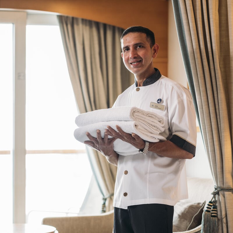 Cabin crew member standing in a cabin area holding neatly folded white towels, with curtains and soft furnishings visible behind.