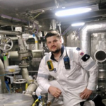 Crew member in engineering overalls standing in a machinery space surrounded by pipes, engines, and industrial equipment.
