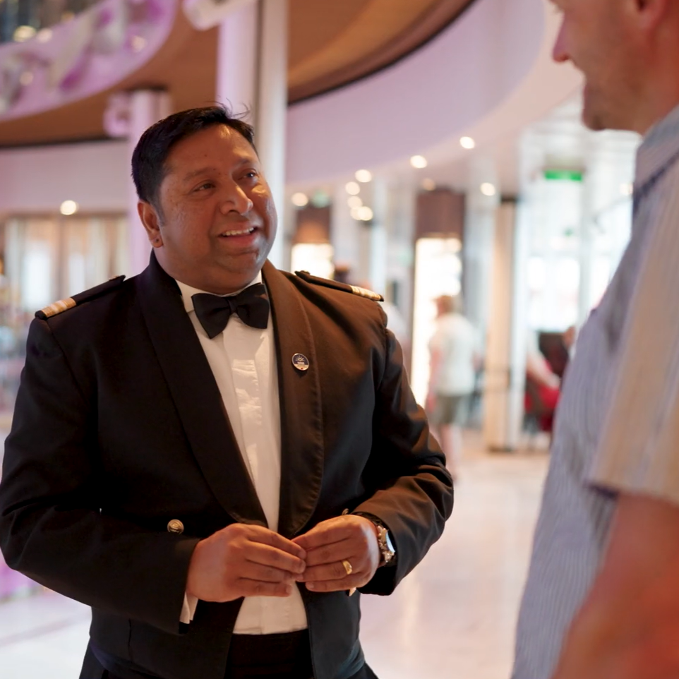 Crew member in formal uniform speaking with another person in a bright dining area, with tables and ambient lighting behind.