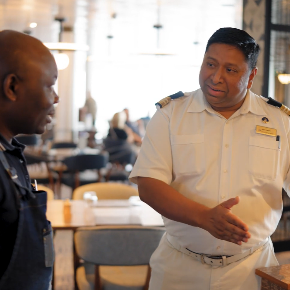 Two uniformed crew members beside a restaurant table, engaged in conversation, with softly lit dining décor in the background