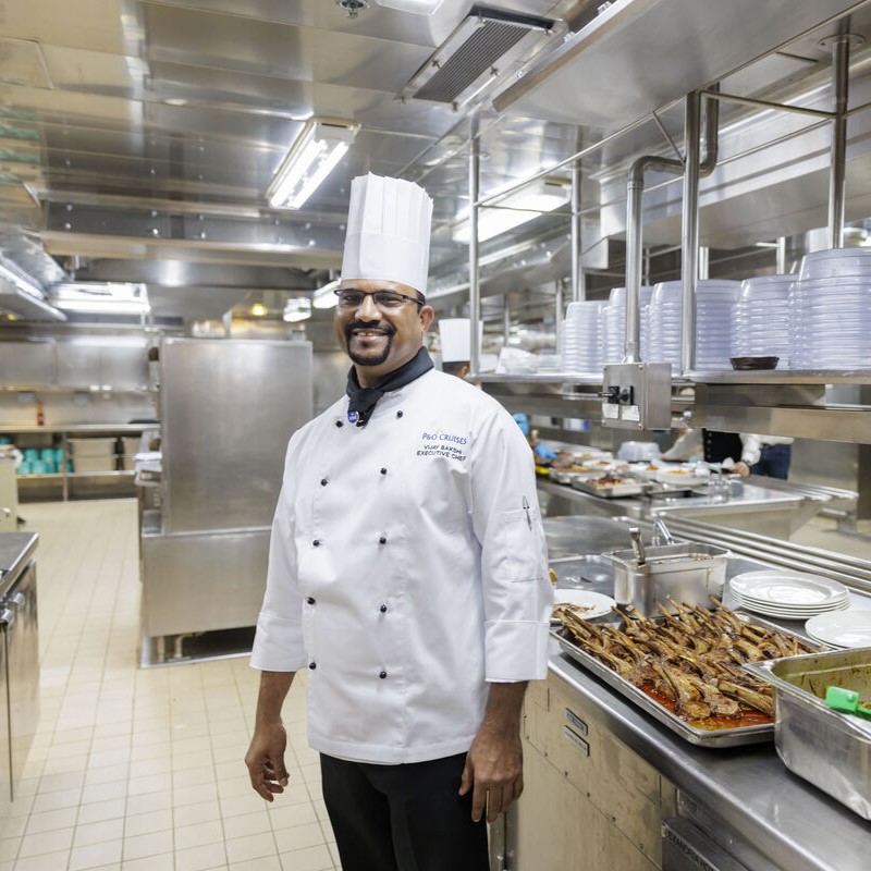 Chef in a white jacket and toque standing alone in a large, modern galley with polished surfaces, overhead racks, and cooking stations.