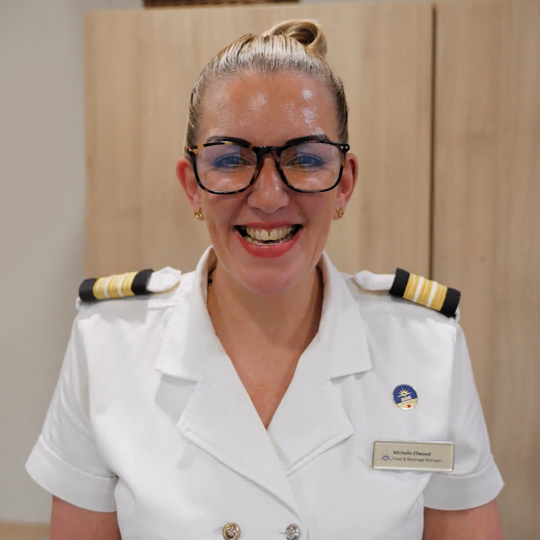 A smiling woman in a white uniform with black and gold epaulettes, wearing glasses and a name badge, stands indoors against a light wooden background—ready for her cruise ship galley job.