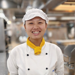A smiling chef wearing a white uniform, white cap, and yellow neckerchief stands in a professional kitchen with stainless steel equipment in the background.
